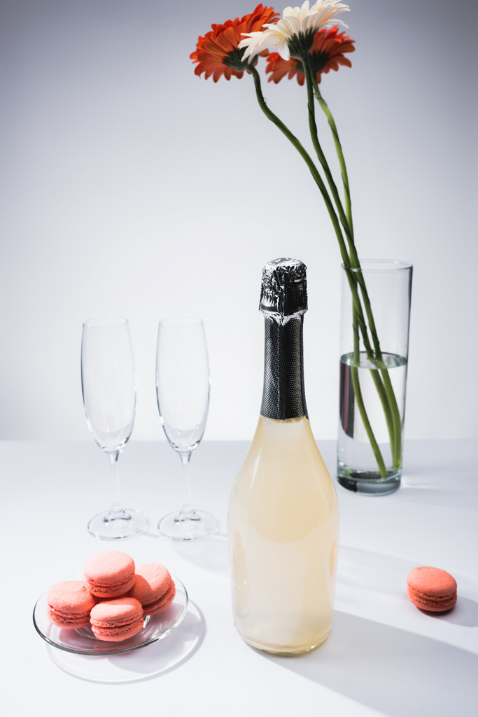 крупним планом вид макарони, порожні окуляри, пляшка шампанського та букет квітів гербери на сірому фоні
 - Фото, зображення