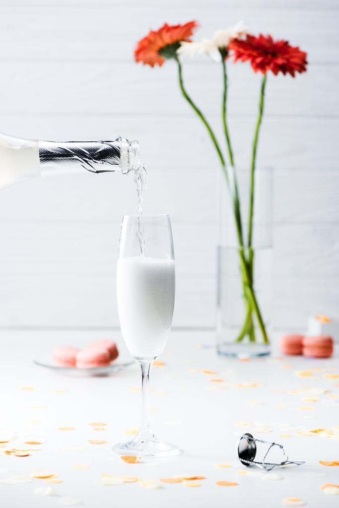 selective focus of pouring champagne into glass process, macarons and bouquet of flowers on grey background - Photo, Image