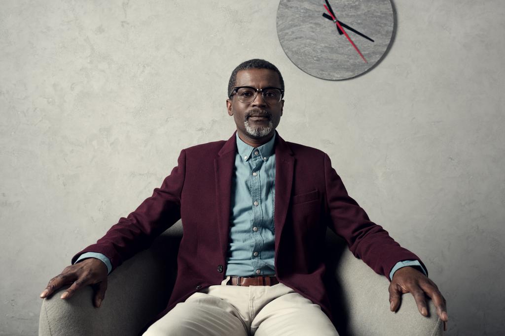 壁の時計との肘掛け椅子に座って自信を持って中央高齢者アフリカ系アメリカ人   - 写真・画像