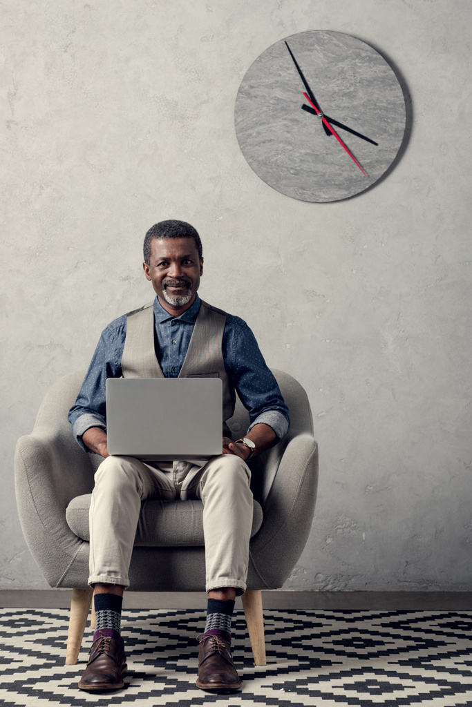 χαμογελώντας Αφρικανικός Αμερικανός επιχειρηματίας χρησιμοποιώντας φορητό υπολογιστή στην πολυθρόνα στο γραφείο με το ρολόι στον τοίχο - Φωτογραφία, εικόνα
