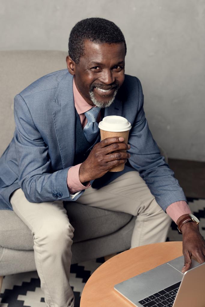 vrolijke volwassen Afrikaanse Amerikaanse man in blauwe jas holding koffiekopje en het gebruik van laptop - Foto, afbeelding