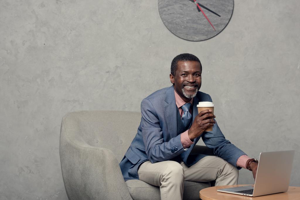 улыбающийся африканский американец в синей куртке с кофе и ноутбуком
  - Фото, изображение