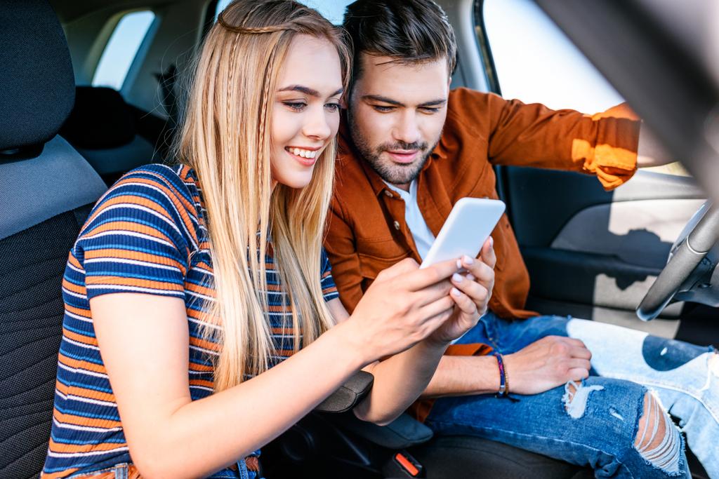 uśmiechnięta Młoda para siedzi w samochodzie i patrząc na ekran smartfona  - Zdjęcie, obraz