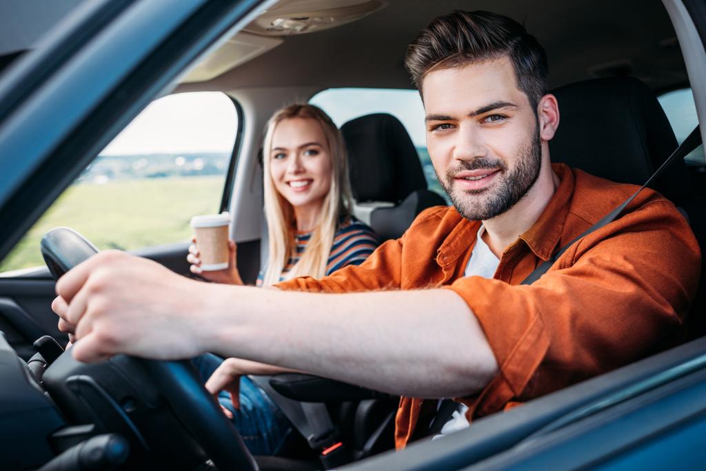 portrait of young man sitting behind car wheel and his girlfriend with paper cup of coffee  - Photo, Image