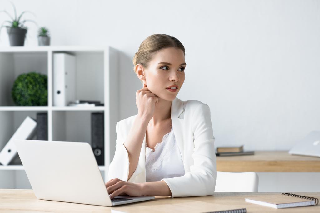 красивая молодая бизнесвумен, работающая с ноутбуком в офисе и отводящая взгляд
 - Фото, изображение