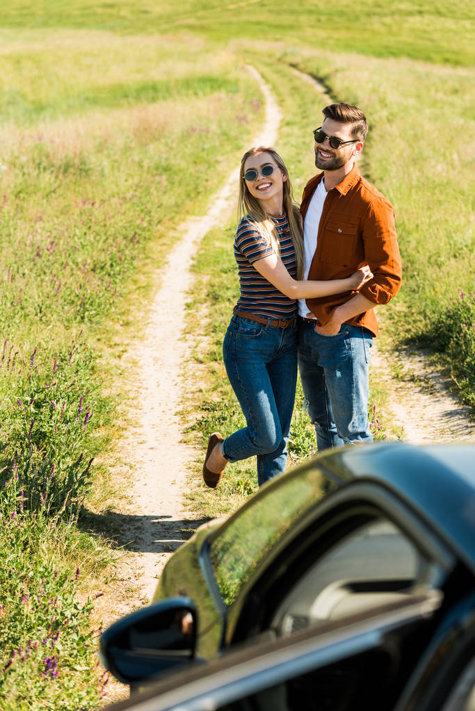 високий кут зору молодої стильної пари в сонцезахисних окулярах, що обіймаються біля автомобіля на сільському лузі
  - Фото, зображення