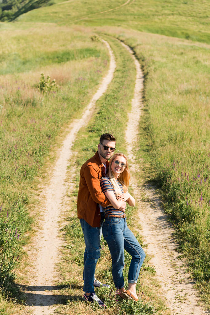 visão de alto ângulo de casal elegante feliz em óculos de sol em pé no prado rural
  - Foto, Imagem