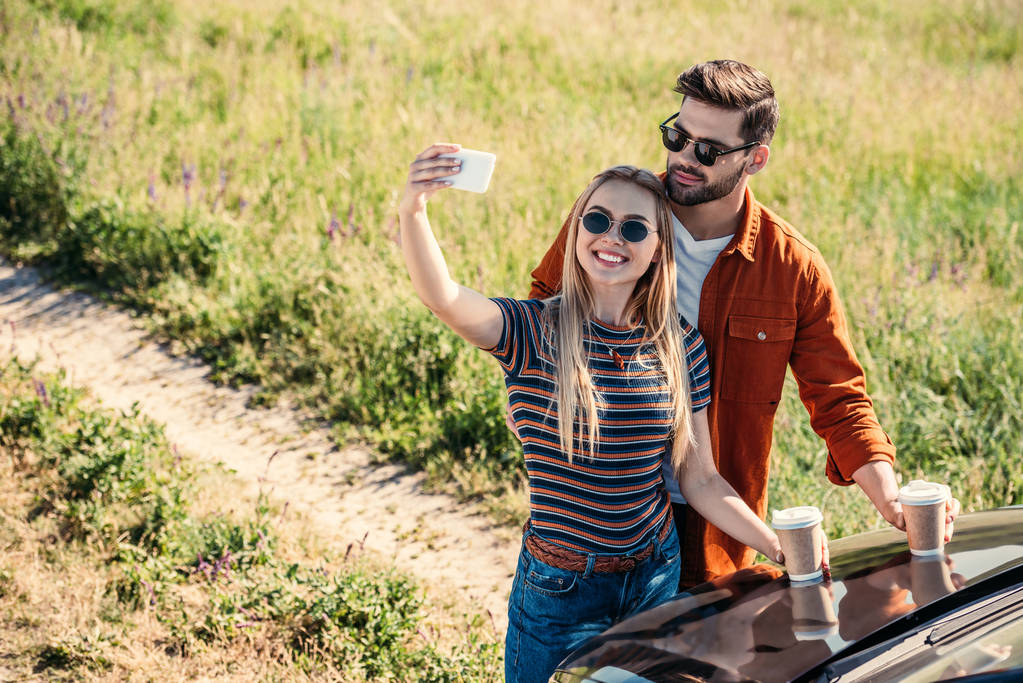 Erhabener Blick auf stylisches Paar mit Sonnenbrille und Kaffeetassen, das in der Nähe des Autos auf der Wiese ein Selfie mit dem Smartphone macht  - Foto, Bild
