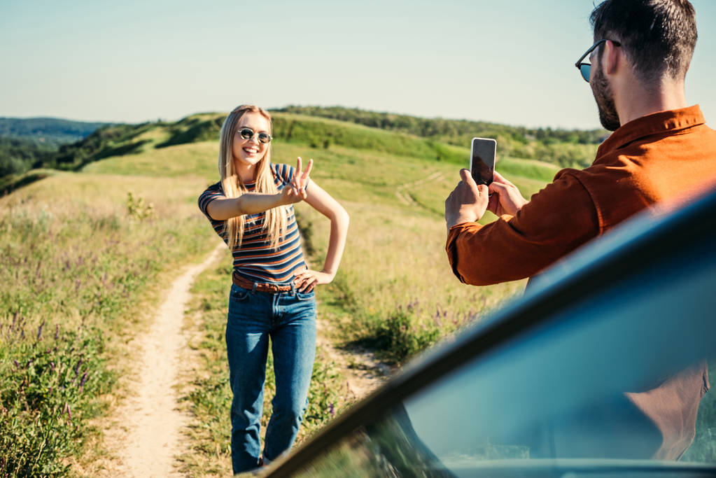 田舎の牧草地の車の近くのピースサインをしているガール フレンドの写真を撮る男  - 写真・画像