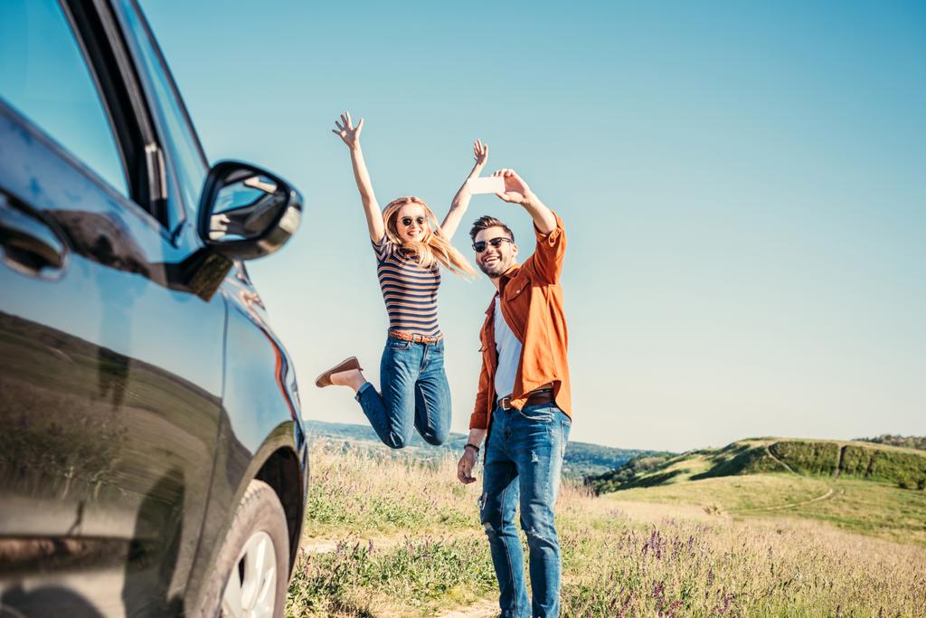 glückliche junge Frau springt mit weit ausgebreiteten Armen, während ihr Freund ein Selfie mit dem Smartphone in der Nähe von Auto im Feld macht  - Foto, Bild