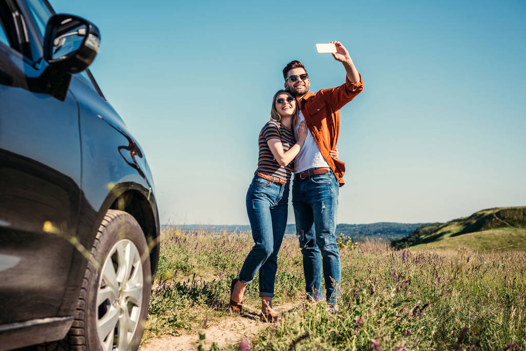 stijlvolle man selfie met vriendin in de buurt van auto op landelijke weide  - Foto, afbeelding