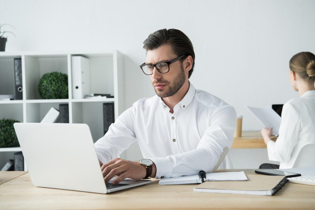 enfocado joven empresario que trabaja con el ordenador portátil en la oficina moderna con colega sentado en el fondo
 - Foto, imagen