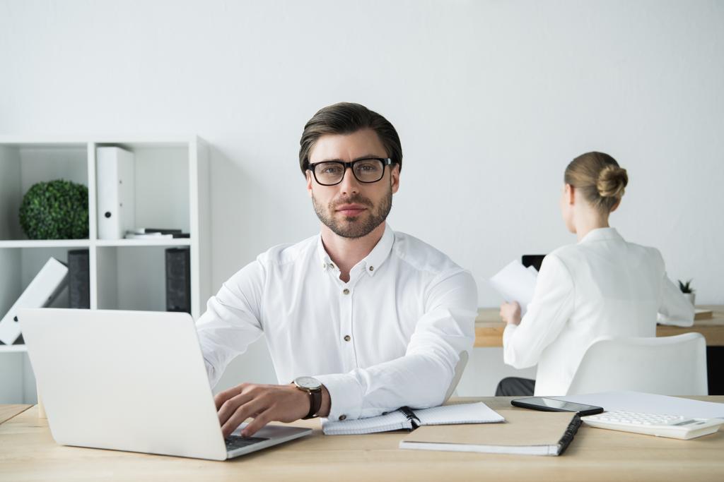 joven hombre de negocios guapo que trabaja con el ordenador portátil en la oficina moderna con colega sentado en el fondo
 - Foto, imagen