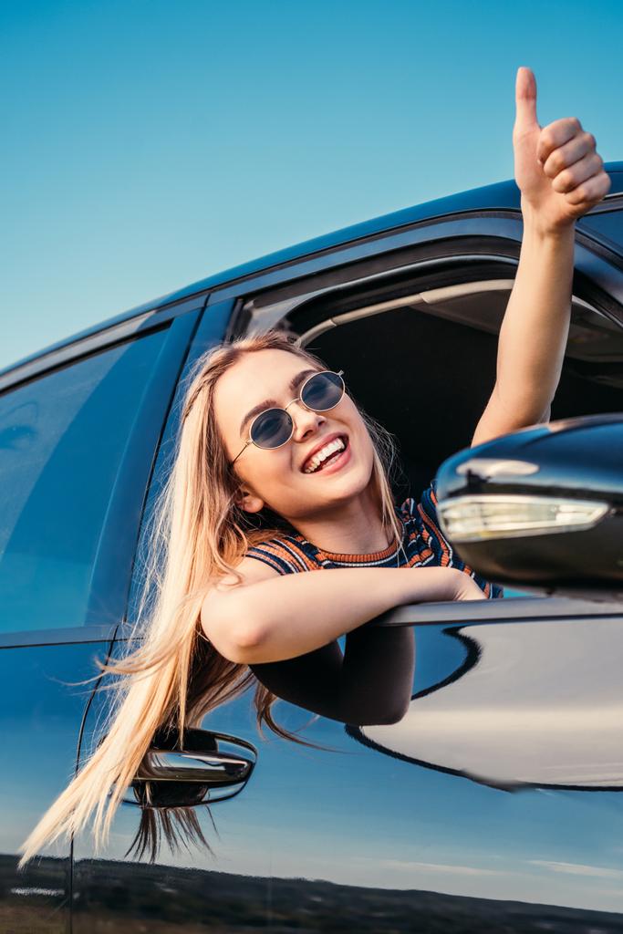 усміхнена стильна жінка в сонцезахисних окулярах виходить з вікна автомобіля і робить жест великого пальця вгору
 - Фото, зображення