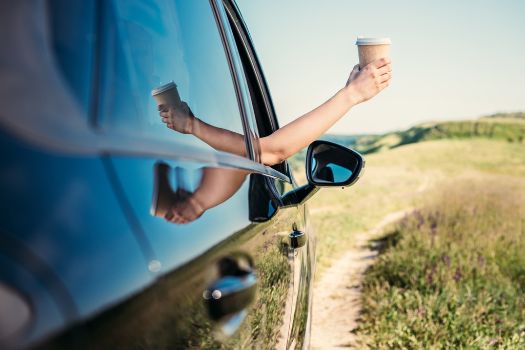 農村フィールドで車の窓からコーヒーの紙コップを持つ手を傾かせる女性のショットをトリミング  - 写真・画像