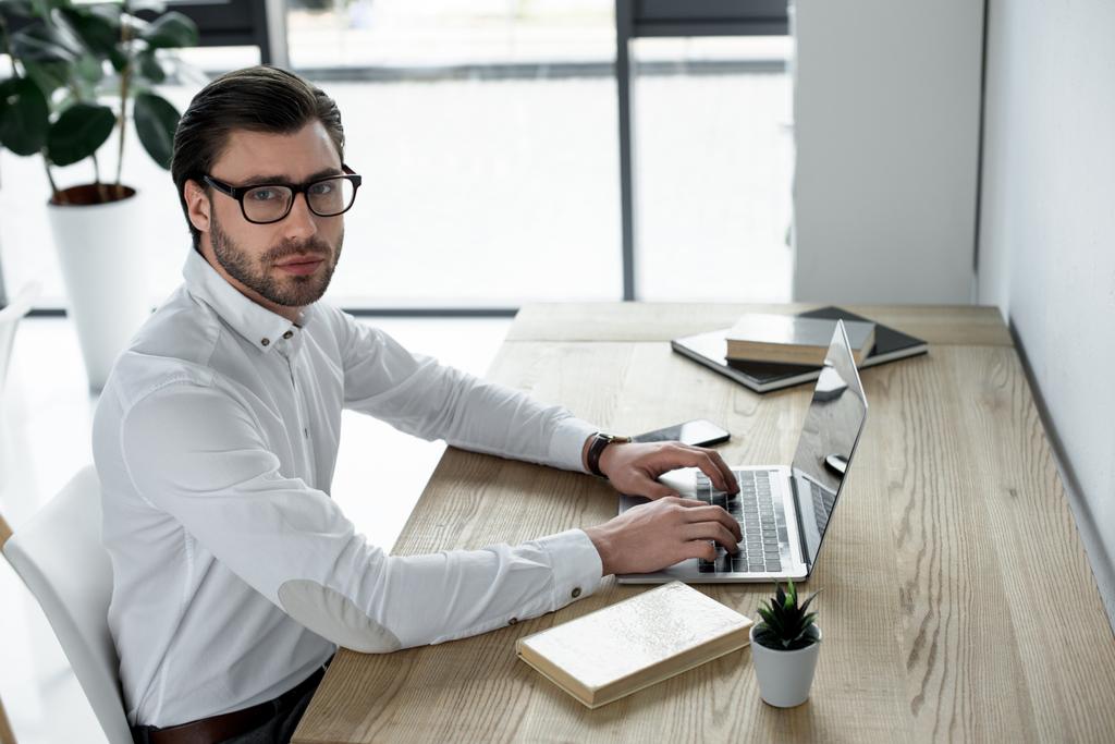 confiante jovem empresário trabalhando com laptop no escritório moderno e olhando para a câmera
 - Foto, Imagem