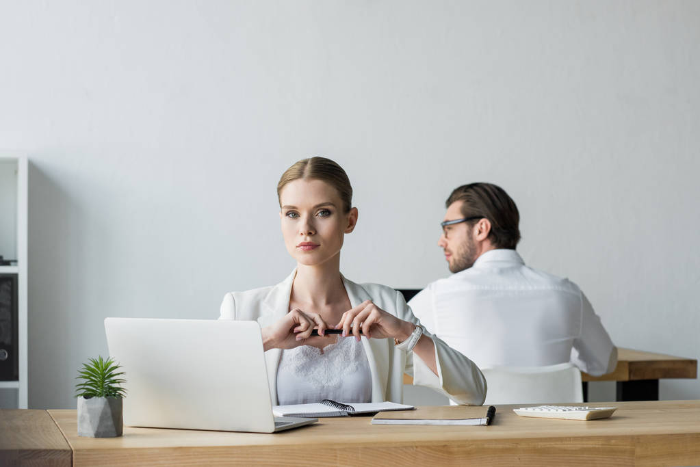 деловая женщина сидит на рабочем месте и смотрит в камеру, пока его коллега работает на заднем плане
 - Фото, изображение