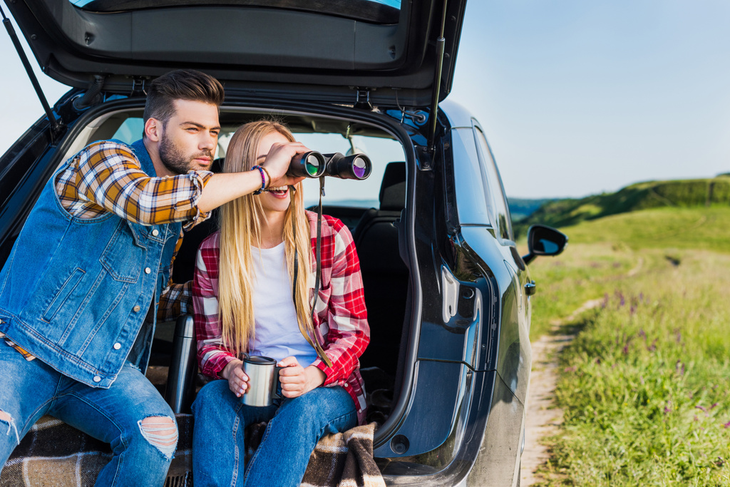Junge männliche Reisende gibt Freundin ein Fernglas, während sie auf dem Kofferraum eines Autos im ländlichen Raum sitzt  - Foto, Bild