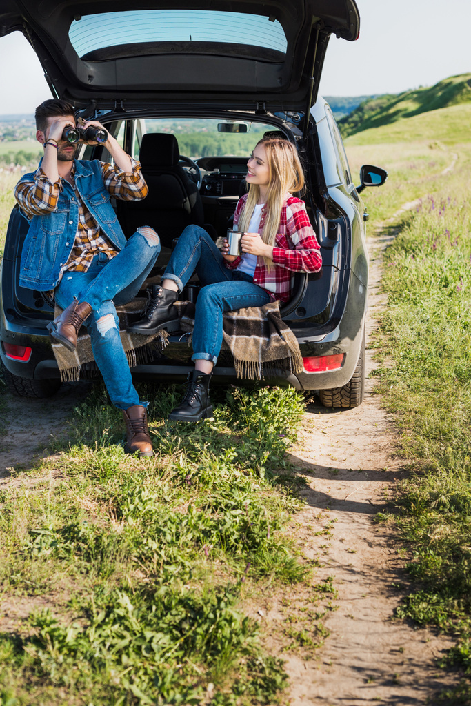 stylish male tourist looking through binoculars while his smiling girlfriend sitting near with coffee cup on car trunk in field - Photo, Image