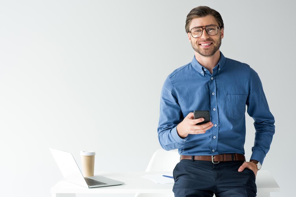 knappe jonge zakenman met smartphone leunend terug op de werkplek geïsoleerd op wit - Foto, afbeelding