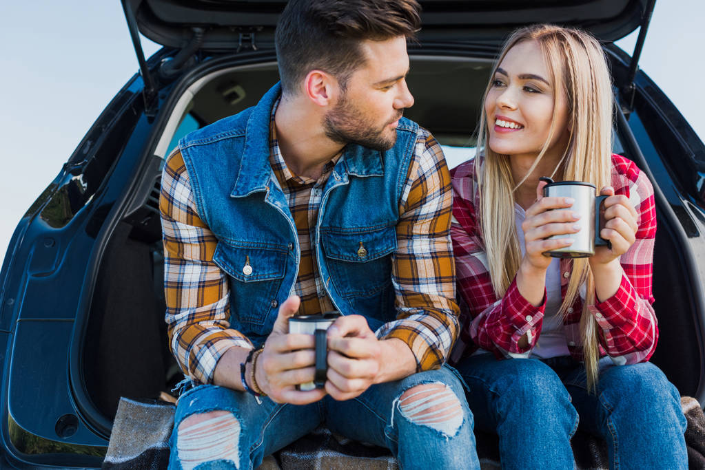 casal de jovens turistas com xícaras de café sentado no porta-malas do carro
 - Foto, Imagem