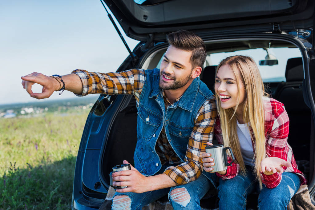 Молодой человек с чашкой кофе указывает рукой на улыбающуюся девушку, сидящую на багажнике машины в сельской местности
  - Фото, изображение