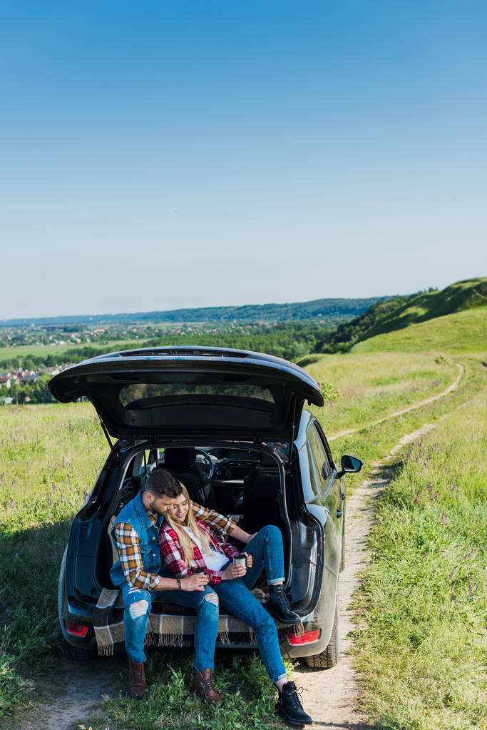 wysoki kąt widzenia kilka stylowych podróżujący z filiżanki kawy siedząc na bagażniku samochodu w wiejskich pola  - Zdjęcie, obraz