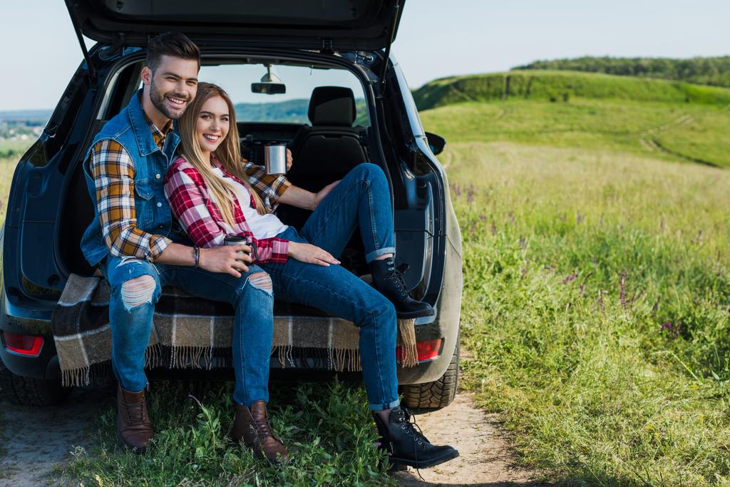 農村フィールドで車のトランクの上に座ってコーヒー カップと笑顔のカップル  - 写真・画像