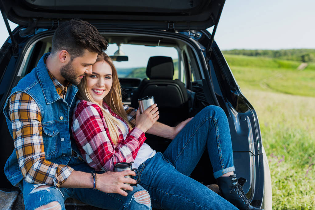 casal de viajantes elegantes com xícaras de café sentado no porta-malas do carro no campo rural
  - Foto, Imagem
