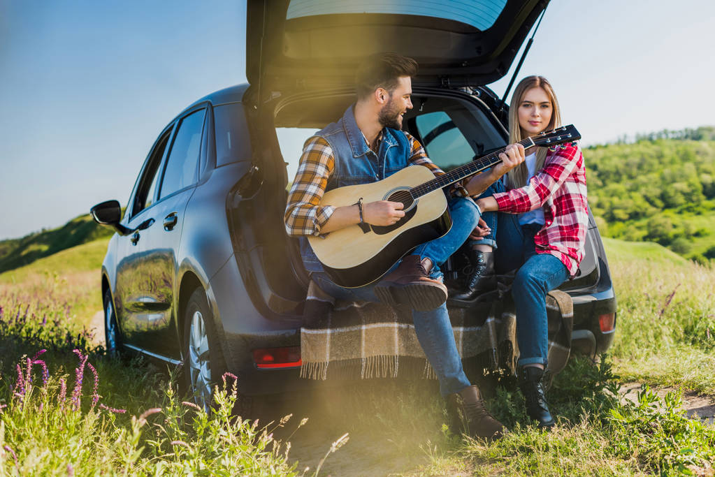 Lächelnder Mann spielt auf Akustikgitarre zu Freundin, die auf Kofferraum im Feld sitzt  - Foto, Bild