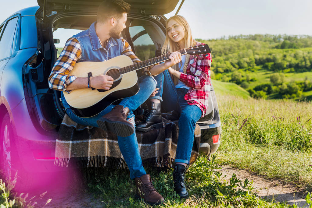 joven viajero masculino jugando en la guitarra acústica a la novia sonriente en el maletero del coche en el campo rural
  - Foto, imagen