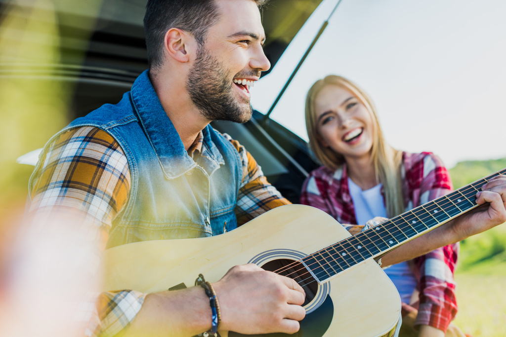 вид сбоку улыбающегося мужчины, играющего на акустической гитаре, в то время как его подруга сидит рядом на багажнике автомобиля
 - Фото, изображение