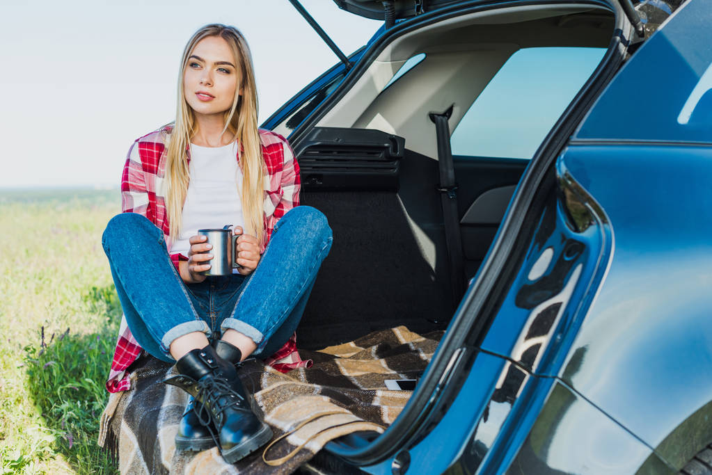 mujer joven sentada en el maletero del coche con taza de café en el campo rural
 - Foto, Imagen