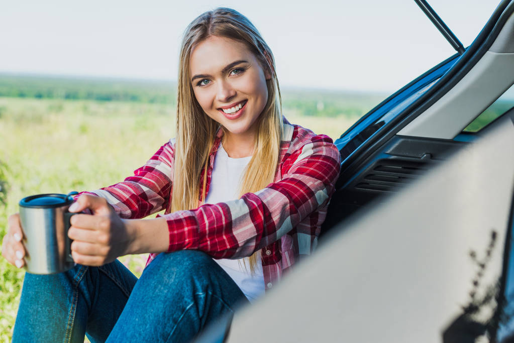 農村フィールド内のコーヒー カップと車のトランクの上に座っている若い女性を笑顔 - 写真・画像