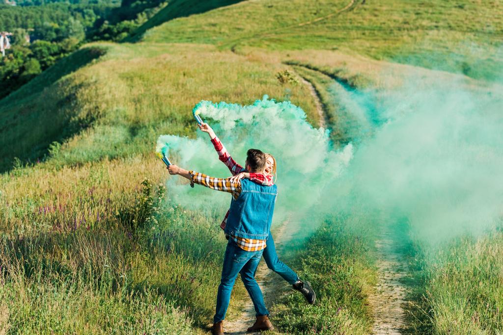 Вид сзади мужчины, держащего в руках подругу и дымовые шашки зеленого цвета на сельской даче
 - Фото, изображение