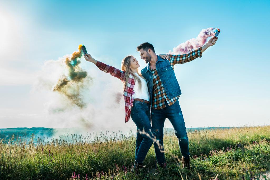 pareja joven mirándose y sosteniendo coloridas bombas de humo en el prado rural
  - Foto, Imagen