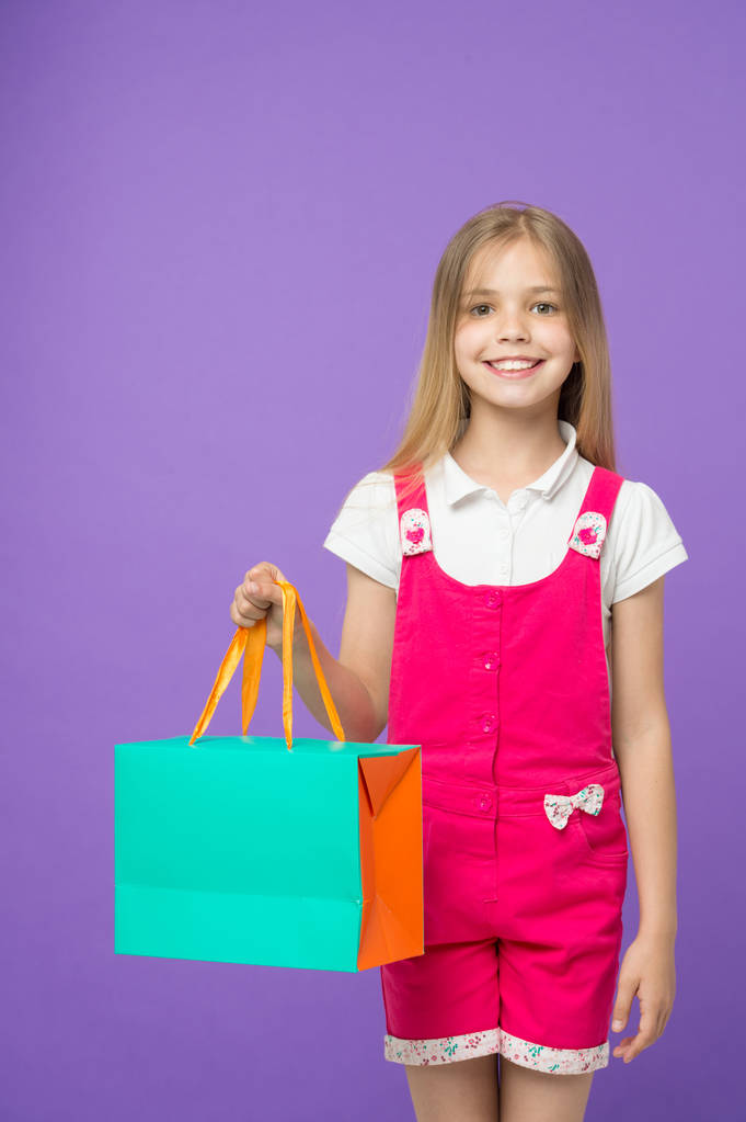 Маленька дитина з сумкою на фіолетовому тлі. Щаслива дівчина посміхається з паперовим мішком. Дитячий покупець у модному комбінезоні. Свята та святкування. Задоволена своїми покупками. продаж і чорна п'ятниця
 - Фото, зображення