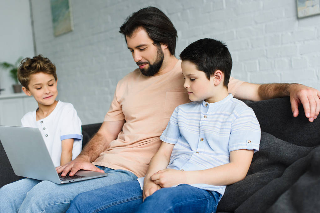 otec a synové poblíž společně pomocí notebooku na pohovce doma - Fotografie, Obrázek
