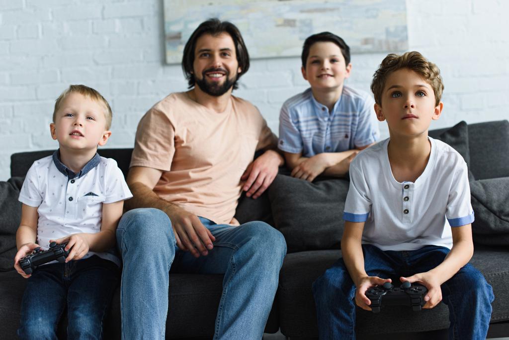 улыбающийся отец смотрит на маленьких сыновей, сидящих на диване и играющих в видеоигры вместе дома
 - Фото, изображение