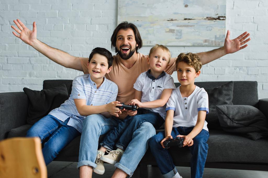 aufgeregter Vater und kleine Söhne sitzen zu Hause auf dem Sofa und spielen gemeinsam Videospiele - Foto, Bild