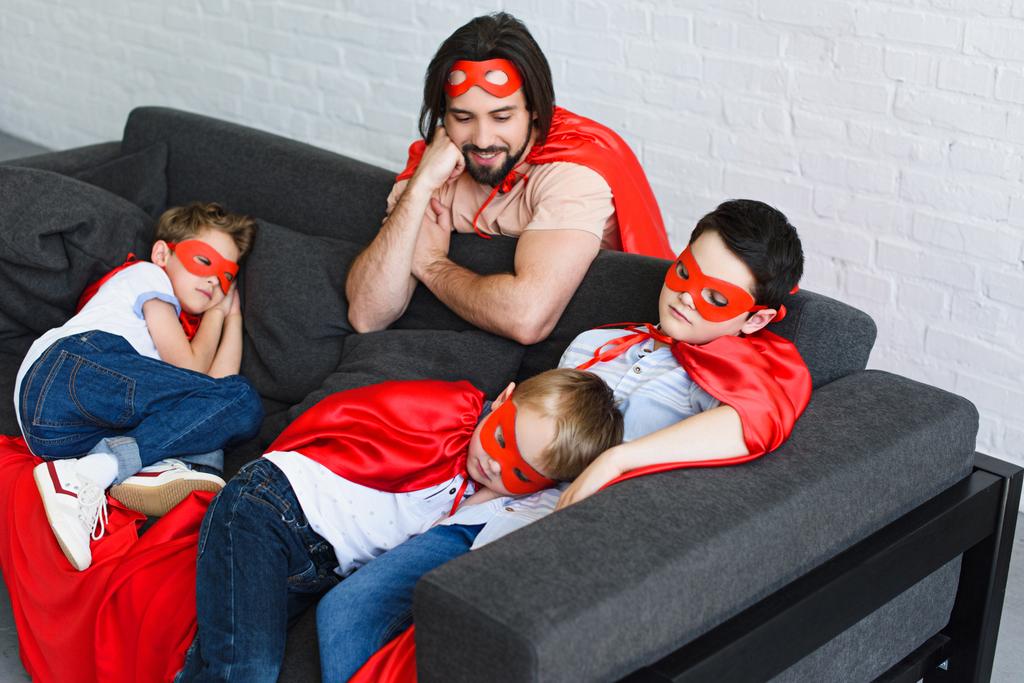 high angle view of happy father looking at sleeping sons in red superhero costumes on sofa at home - Photo, Image