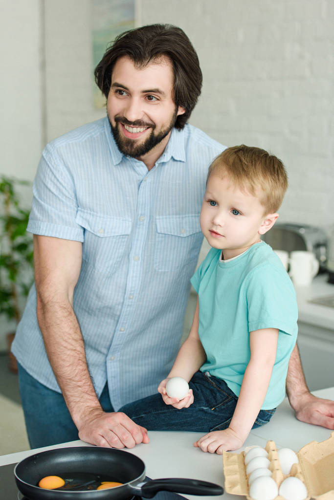 портрет улыбающегося отца и маленького сына с сырым яйцом в руке на кухне дома
 - Фото, изображение