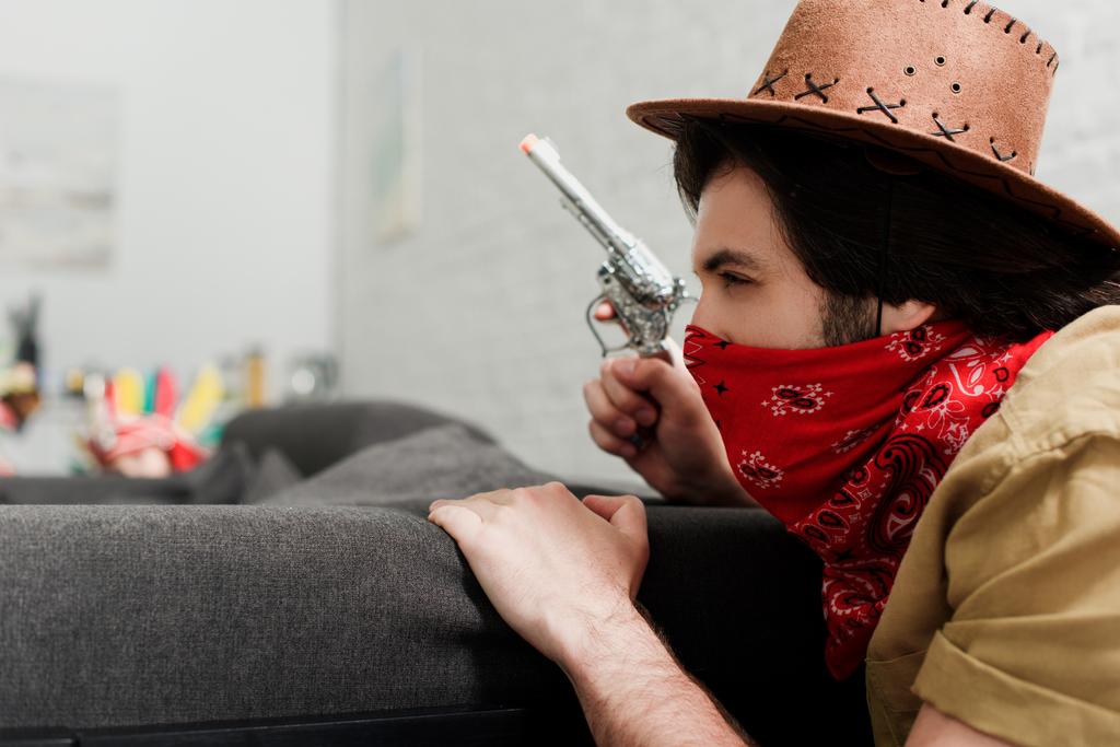 Widok z boku człowieka w czerwonym i kowbojski kapelusz z pistolet zabawka na kanapie w domu - Zdjęcie, obraz