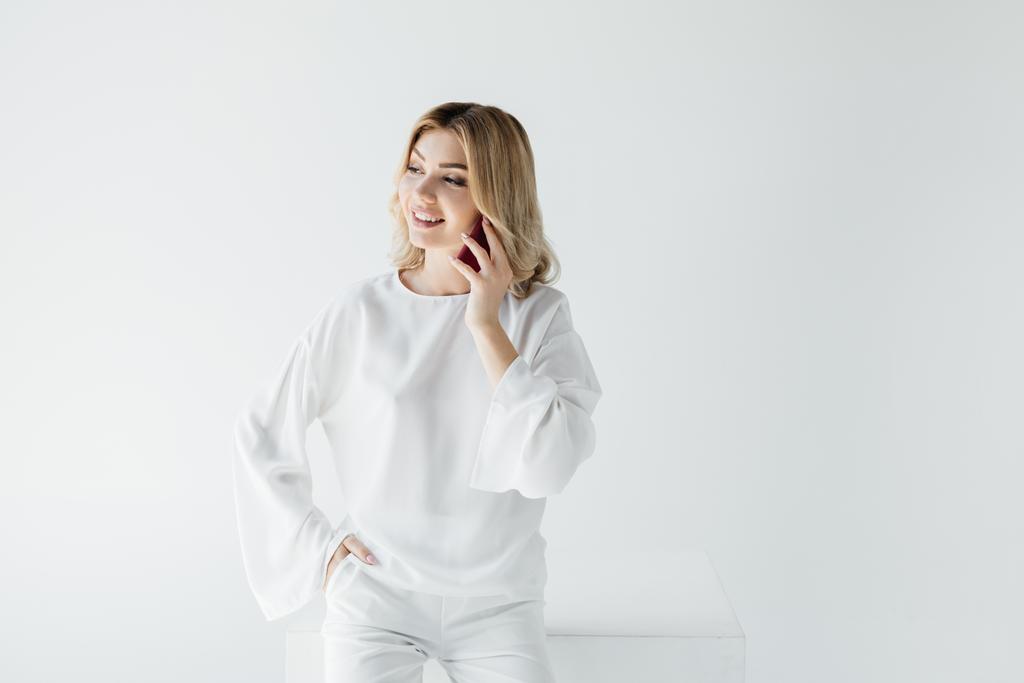 Portret van lachende blonde vrouw in witte kleding praten op smartphone geïsoleerd op grijs - Foto, afbeelding