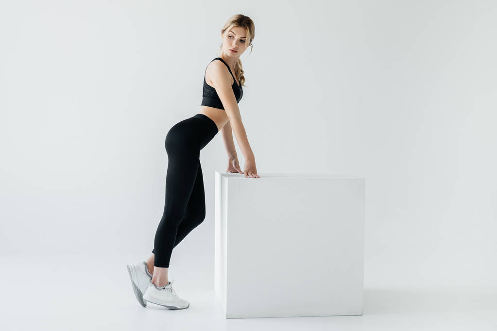 side view of sportive woman in black sportswear standing near white cube on grey background - Photo, Image
