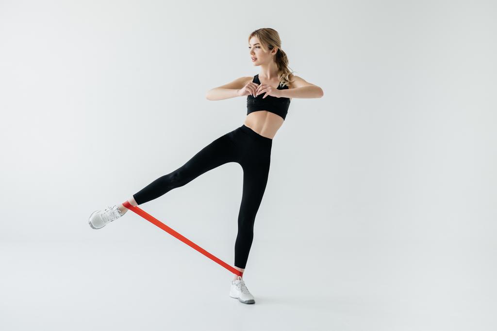 junge athletische Frau trainiert mit Gummiband an Beinen isoliert auf grau - Foto, Bild