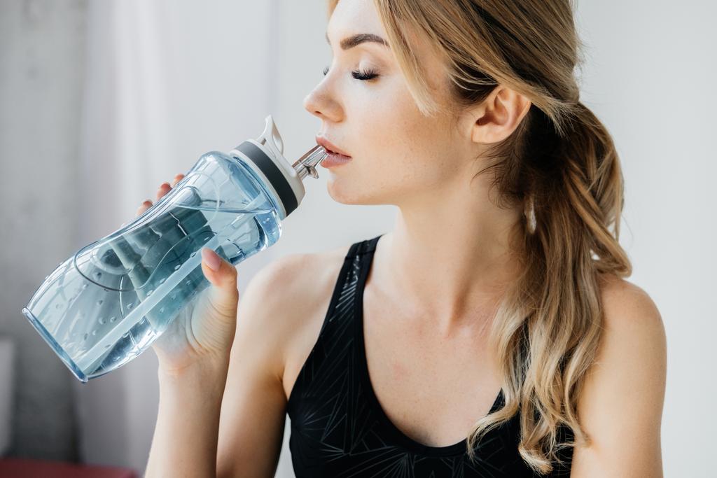 Seitenansicht athletische Frau trinkt Wasser aus sportlicher Wasserflasche vor grauem Hintergrund - Foto, Bild