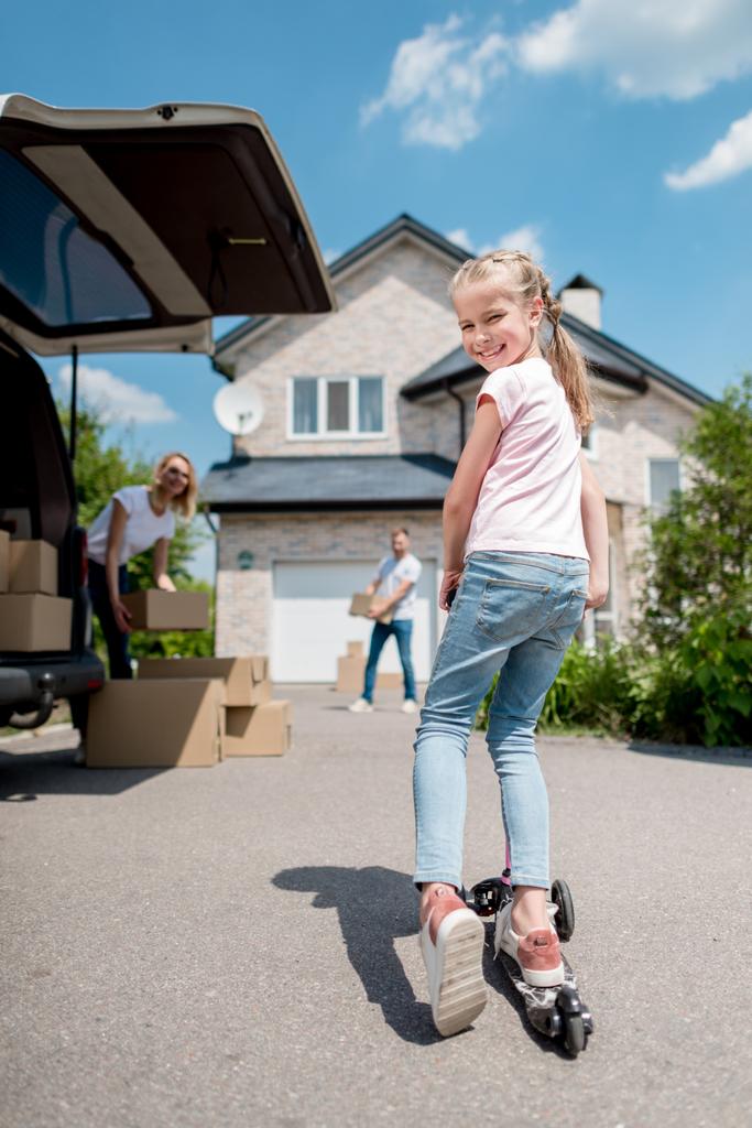 Gelukkig kleine jongen rijden op kick scooter en haar ouders uitpakken van kartonnen dozen voor verhuizing naar nieuw huis  - Foto, afbeelding