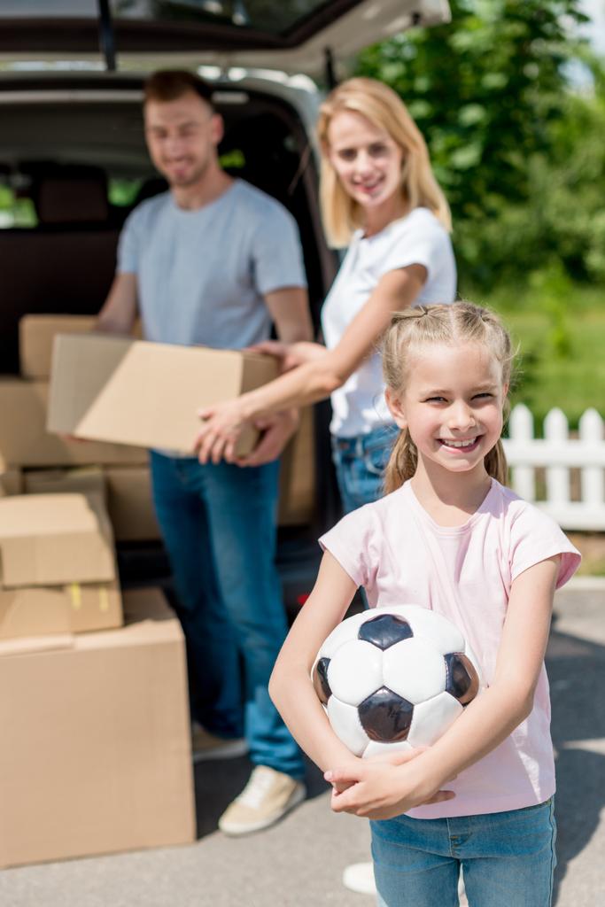 glückliches kleines Kind mit Fußballball, während ihre Eltern Kartons für den Umzug auspacken  - Foto, Bild