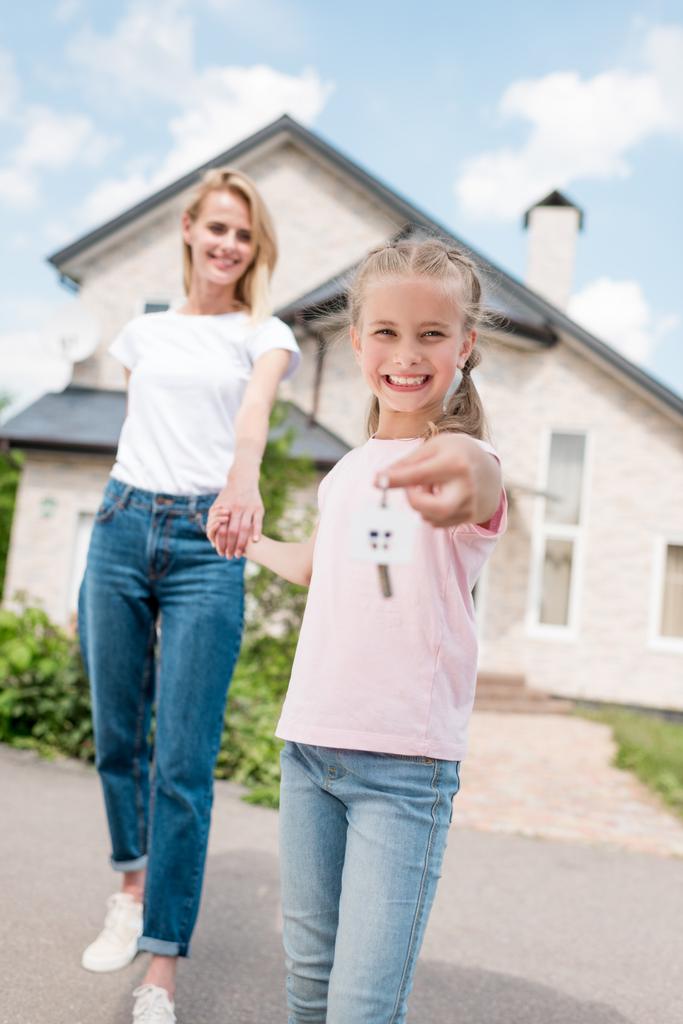 glücklich lächelndes Kind zeigt Schlüssel mit Schmuckstück und hält Hand der Mutter vor dem neuen Haus  - Foto, Bild